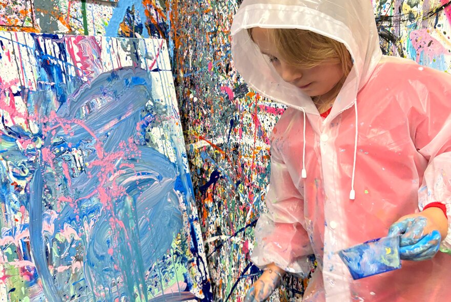 A child wearing a plastic coat, painting a canvas that rests against a wall splattered with paint.