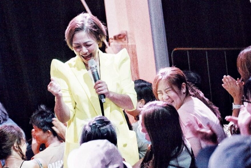 A comedy holding a microphone standing next to seated women.