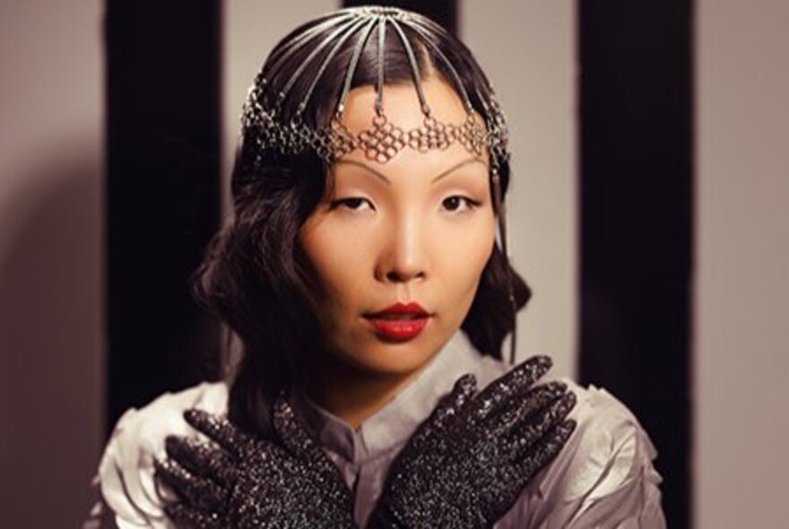 Singer Dami Im, wearing a decorative headpiece on the top of her head, and holding her long sparkly gloves up under her chin.