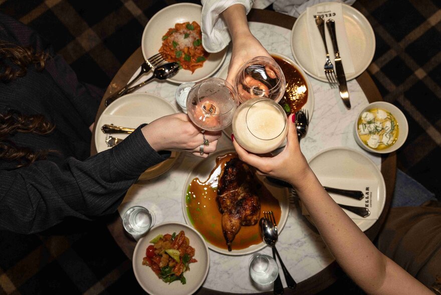 Three hands holding cocktails over a round table filled with plates, cutlery and share dishes, all seen from above.