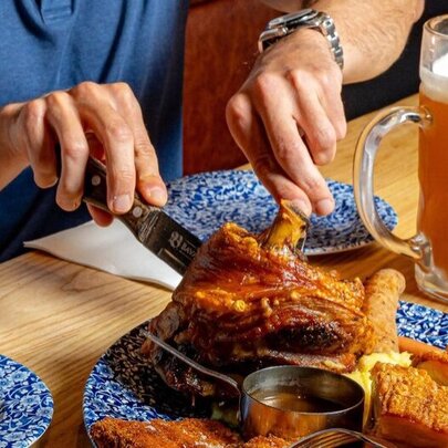 Hands tucking into a huge plate of pork knuckles with a large glass of beer.