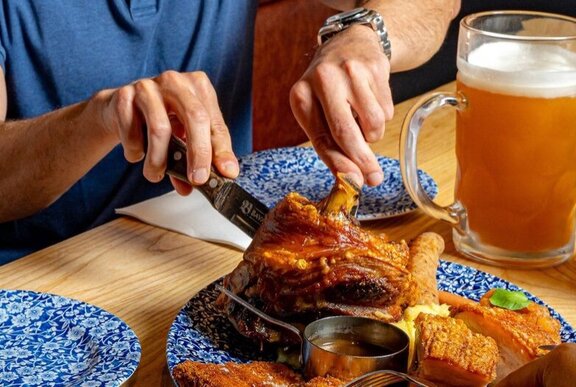 Hands tucking into a huge plate of pork knuckles with a large glass of beer.