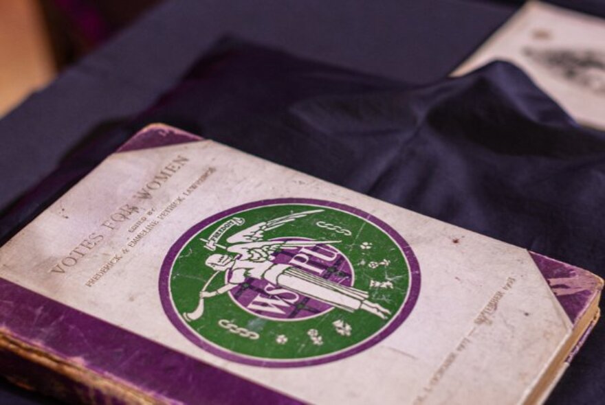 A vintage copy of a book titled, Votes For Women, from the WSPU, resting on a blue silk cloth.