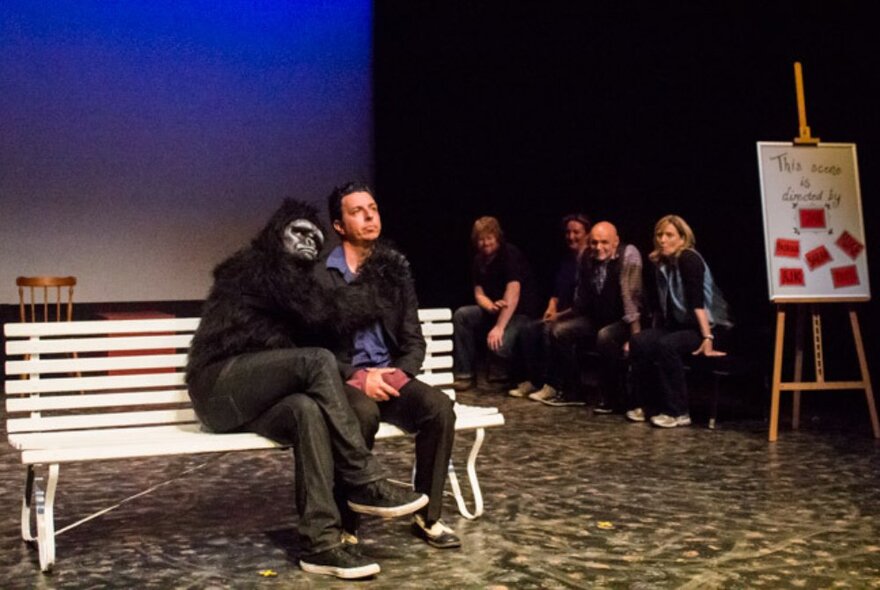 Actors on a stage, four in the background behind an easel, and a man in the foreground on a park bench seated next to someone wearing a gorilla costume.