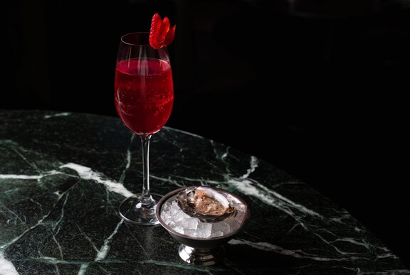 A vibrant red\ spritz alongside a single oyster served on a marble counter.
