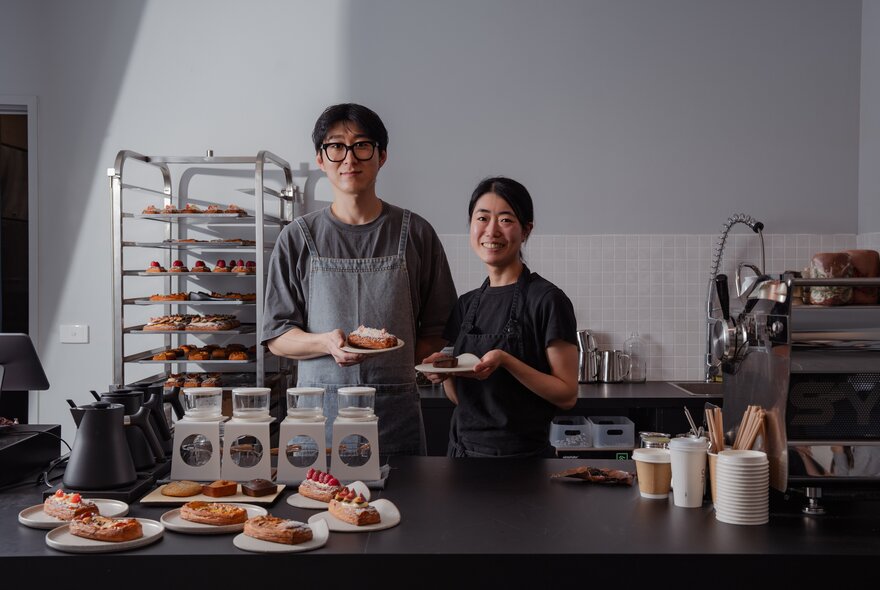 Two bakers holding plates with pastries behind a counter with products and pastries.