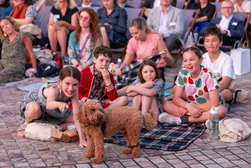 Young kids sitting cross-legged on a picnic blanket on a paved surface, with a fluffy brown dog.