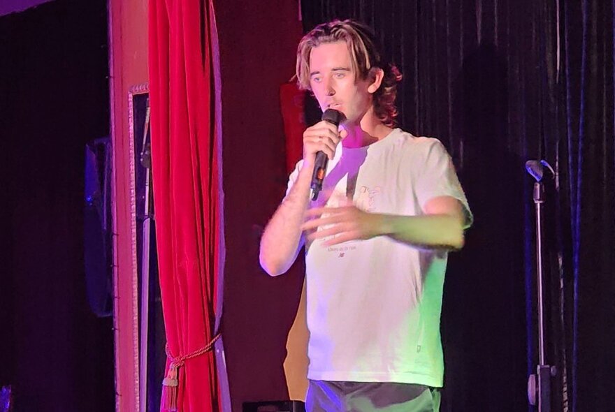 A male stand-up comedian holding a microphone and performing on a stage.