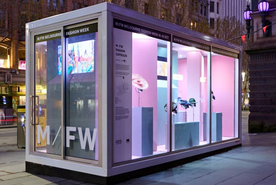 A Melbourne Fashion Week outdoor fashion capsule installation of fashion displayed in a  large glass rectangular case, situated in a public space.