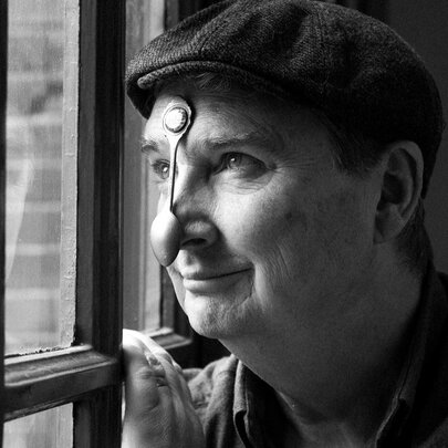 Black and white photograph of comedian Damien Callinan wearing a peaked cap and a teaspoon stuck to his nose as he looks out a window.