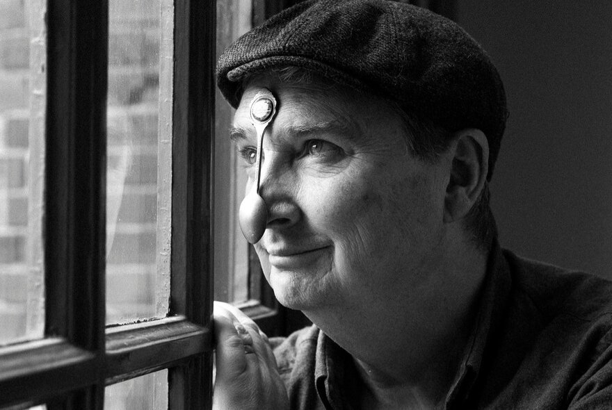 Black and white photograph of comedian Damien Callinan wearing a peaked cap and a teaspoon stuck to his nose as he looks out a window.