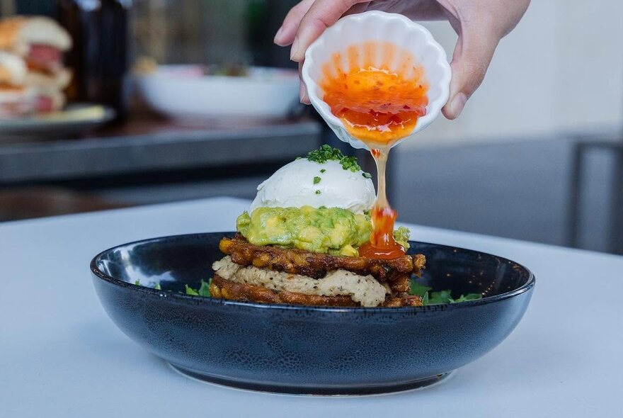 A hand pouring a chili sauce from a small dish over a cooked and loaded egg breakfast, served in a shallow black bowl.