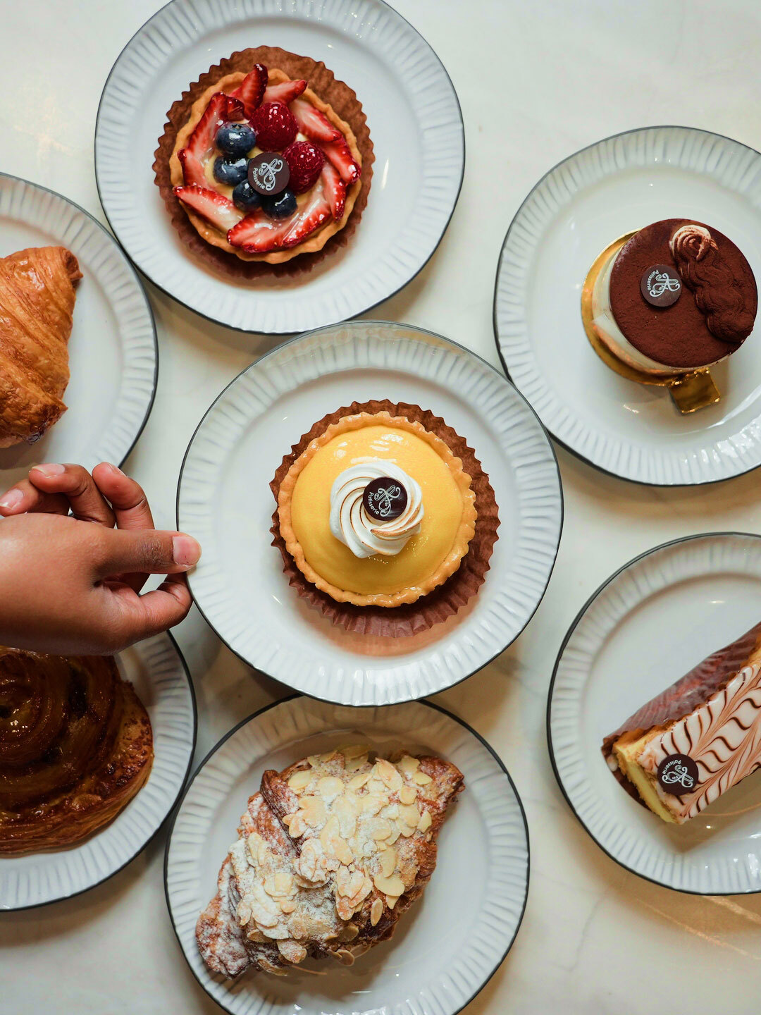 A hand picking up a plate with a tart on it, surrounded by a variety of French desserts.