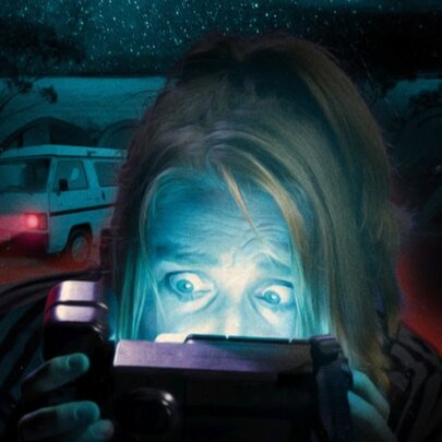 Coloured image of a woman behind a camcorder looking terrified, a campervan in the the background. 