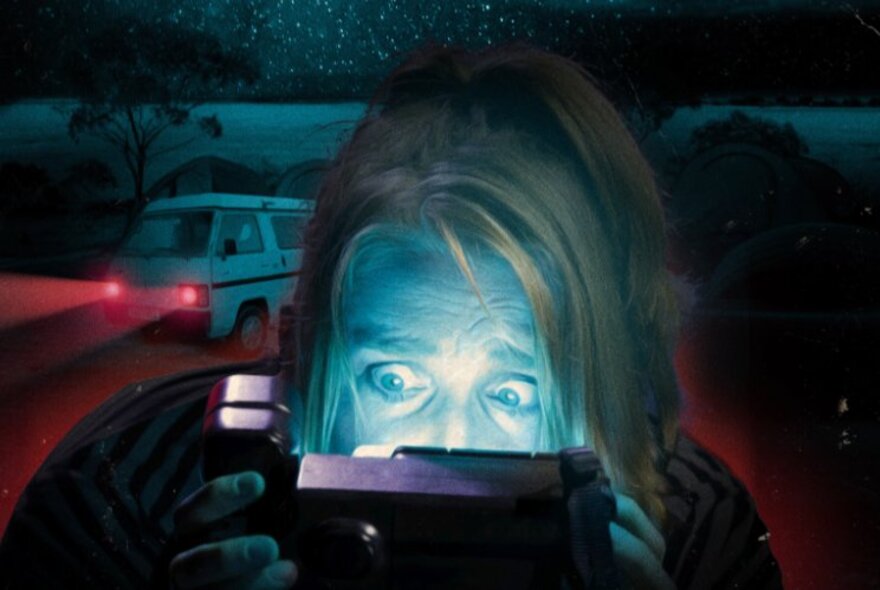 Coloured image of a woman behind a camcorder looking terrified, a campervan in the the background. 