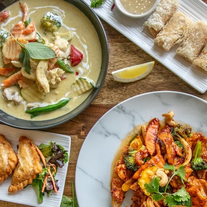 Top down view of plates of Thai food including a green curry, and a stir fry, on a wooden table.