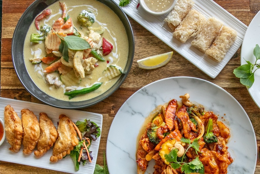 Top down view of plates of Thai food including a green curry, and a stir fry, on a wooden table.