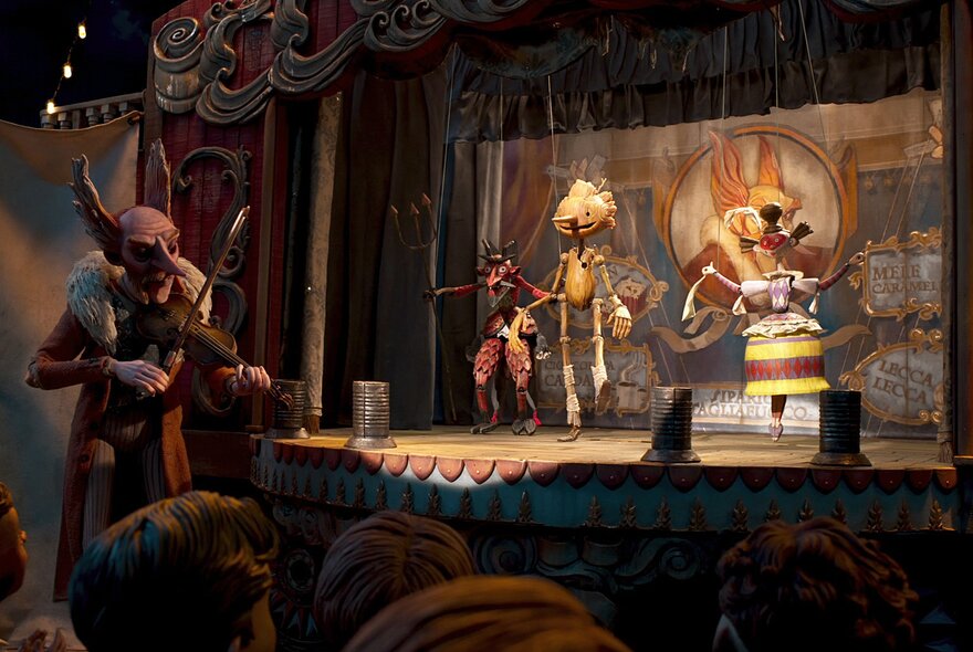 A still image from a film showing three marionettes on a stage with a devil-like creature at the side of the stage playing a violin.