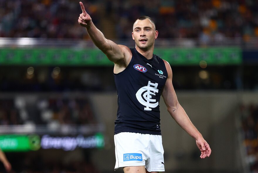 A Carlton AFL player celebrating on the field, with his arm raised and finger pointing skywards, blurred fans in the stadium behind him.