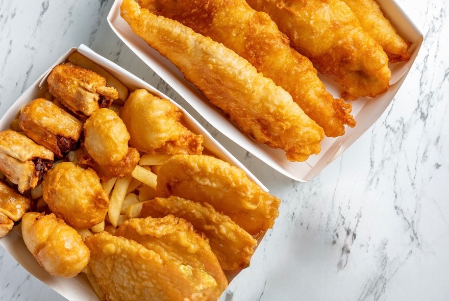 Looking down on two paper trays from a fish and chip shop with fish, dim-sims and potato cakes. 