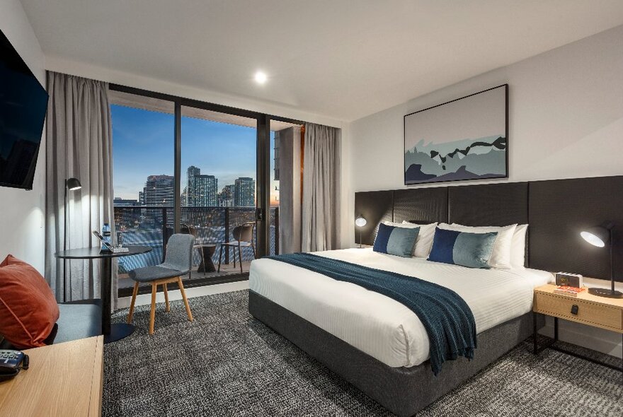 A bedroom at Quest NewQuay with a king size bed, bedside tables, a small table by the window, grey carpet and views of the Melbourne city skyline.