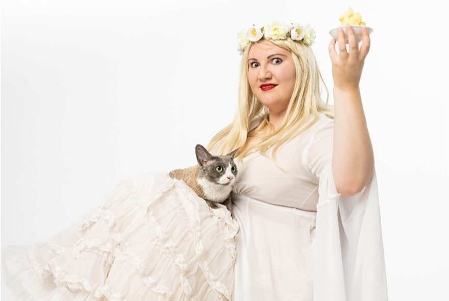 A woman in a white gown with a floral garland around her head holding a cat in the folds of her dress and a small plate of food in her raised hand. 