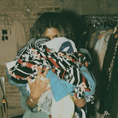Person's face hidden behind the pile of clothing they're holding, next to racks of clothing.