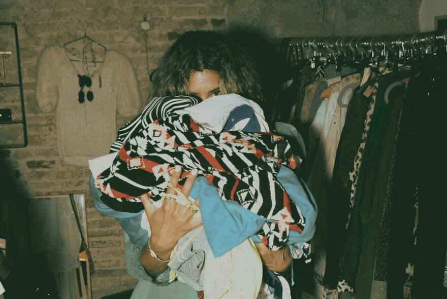 Person's face hidden behind the pile of clothing they're holding, next to racks of clothing.