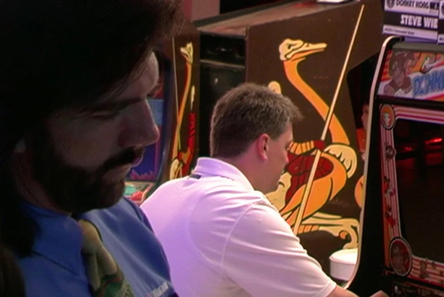 Two men in an arcade setting playing retro arcade games. 