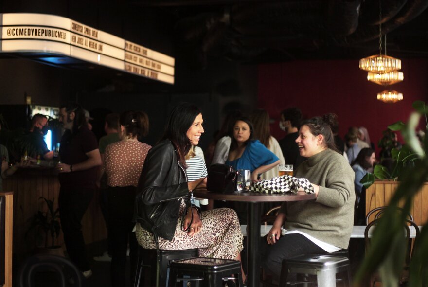 An indoor bar where two women are seated at a round high bar table talking, with other patrons in the background.