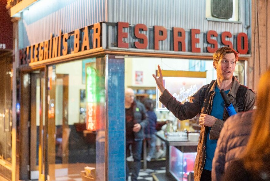 A tour guide talking to people standing in front of Pellegrini's Bar, at night.