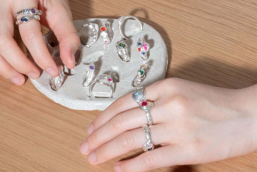Hands covered in silver rings and a mat with other rings on display. 