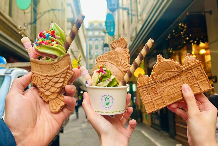 Three hands holding up waffles shaped like fish and one shaped like Flinders Street Station, some topped with soft-serve ice-cream, against a backdrop of Degraves Street in the city.