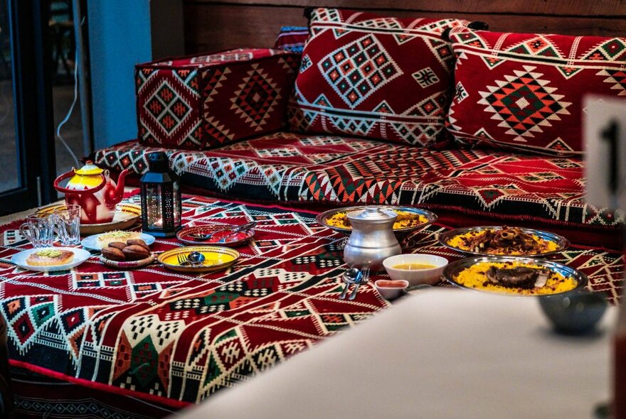 Silver platters of Yemeni food presented on a low table with matching seating, including bowls of rice and meat, with glasses and table lantern.