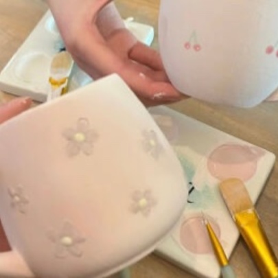 Hands holding white ceramic mugs that display painted designs, with brushes and paints nearby, as part of a creative workshop. 