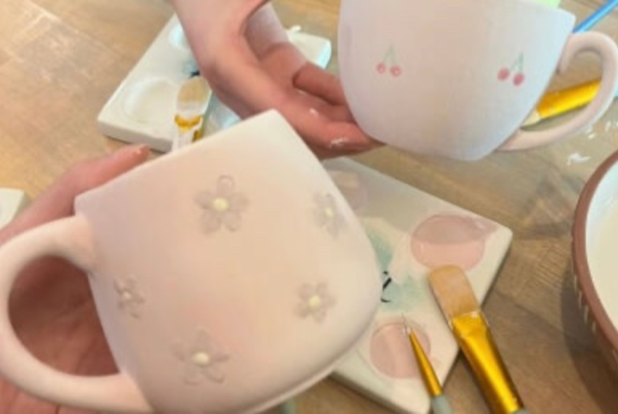 Hands holding white ceramic mugs that display painted designs, with brushes and paints nearby, as part of a creative workshop. 