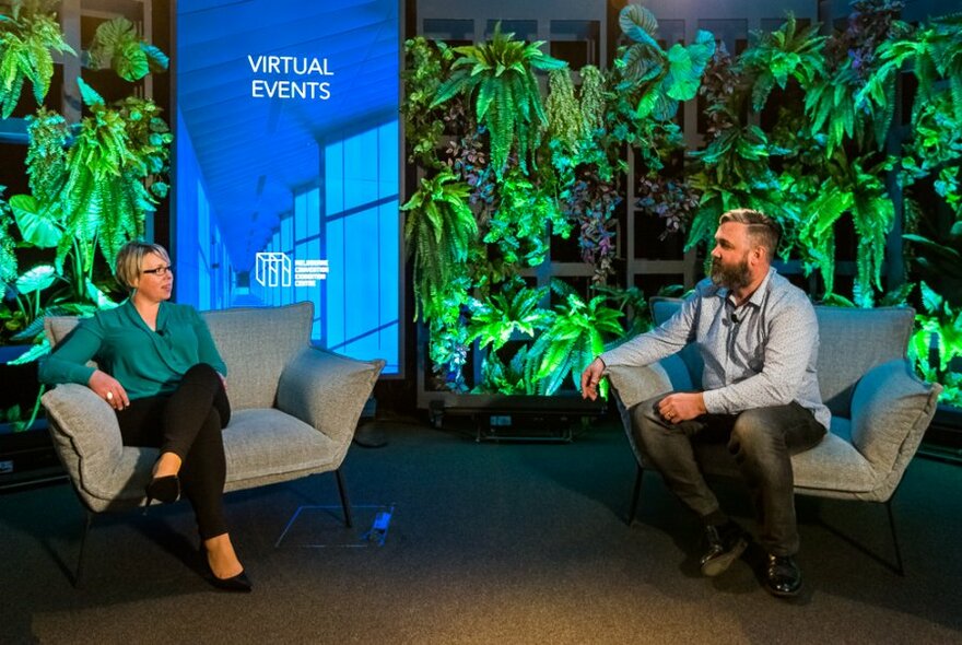 Two people in armchairs talking to each other with plants in the background.