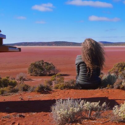 Woman seated on in a red desert landscape, with her back to the viewer, next to a piano under blue skies.