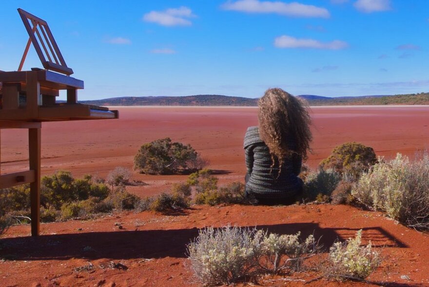 Woman seated on in a red desert landscape, with her back to the viewer, next to a piano under blue skies.