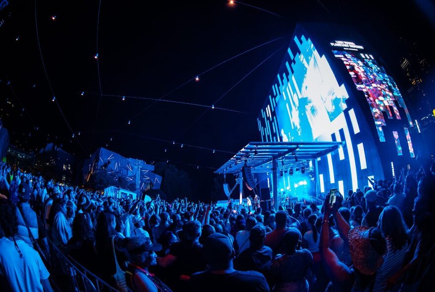 A large audience watching a live music gig on stage at Fed Square at night.