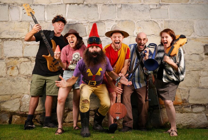 Gardener Costa dressed as a garden gnome, bending forward excitedly with other performers holding instruments and smiling.