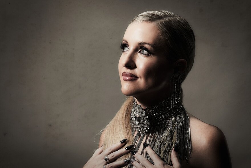 Woman with hands at her throat wearing elaborate necklace and sweeping blonde hair.