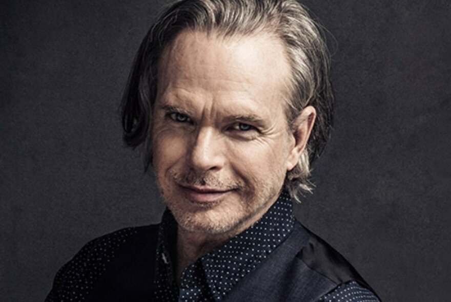 A head shot of singer Rick Price in a black shirt with small white polka dots, smiling warmly. 