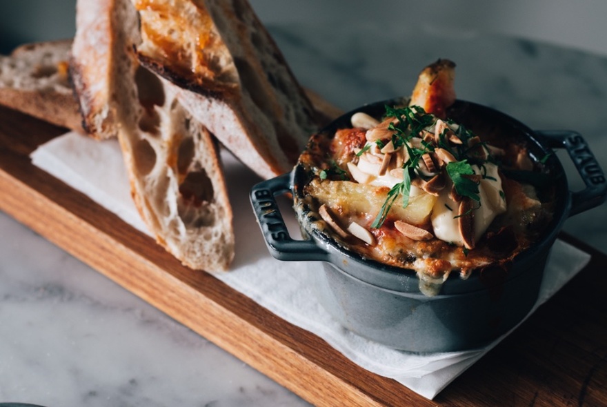 A wooden platter with bread and a mini pot of casserole for one.