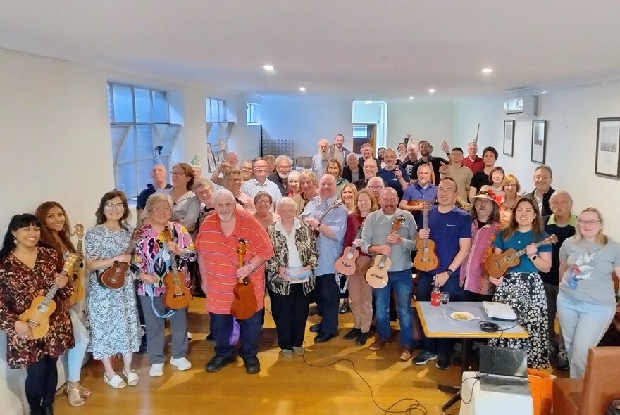 A large group of people all holding ukeleles as they pose for a photo in a white-walled room.