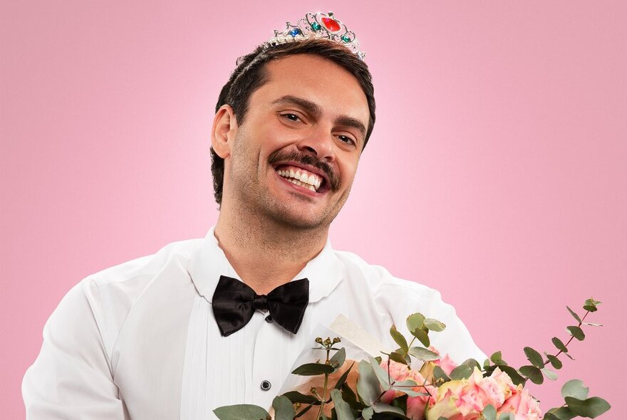 Comic smiling wearing a tiara on his head and black bow tie, holding a bunch of flowers.