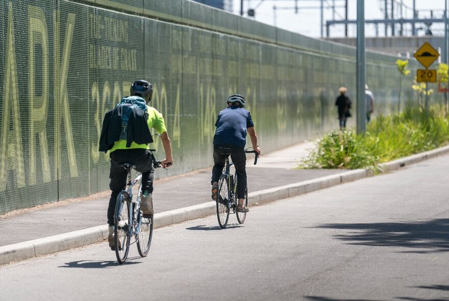 Cyclists riding their bikes on a road past a high green fence.