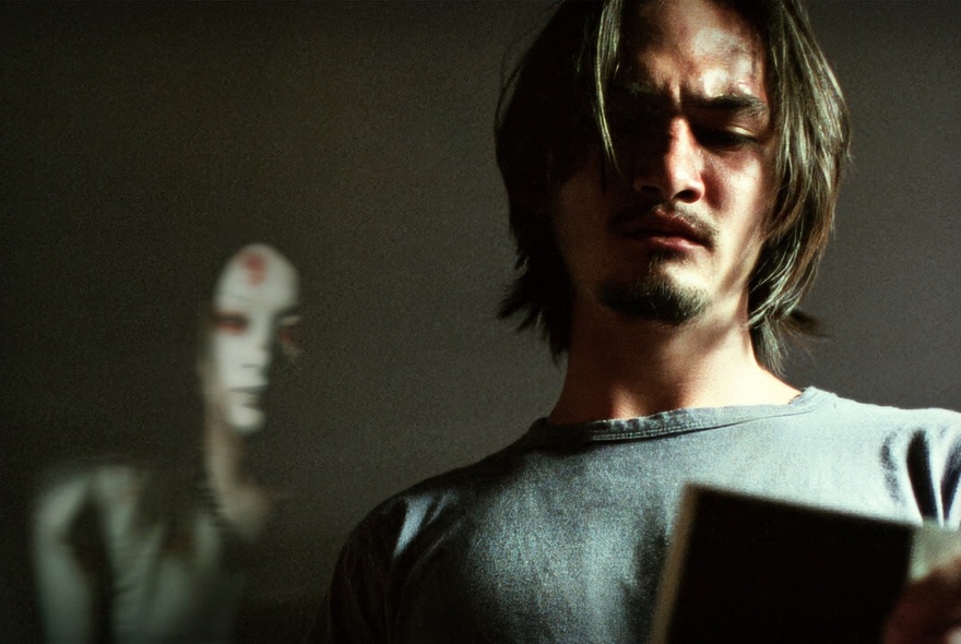 A still image from the movie Shutter, showing a man with a pained expression looking downwards and a ghostly female figure behind him. 