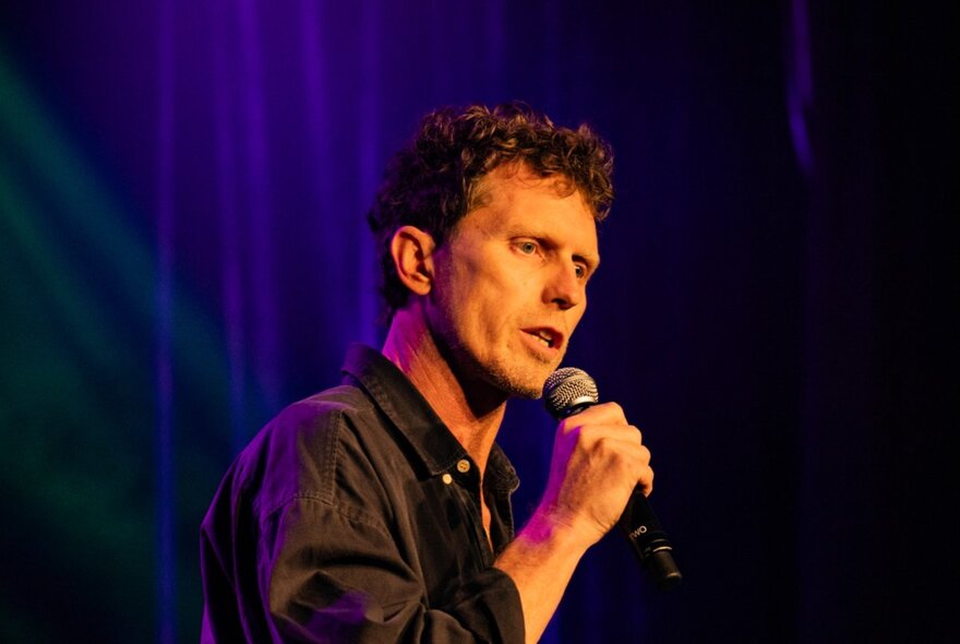 Australian comedian, David Quirk, on stage holding a microphone; a dark blue velvet curtain behind him.