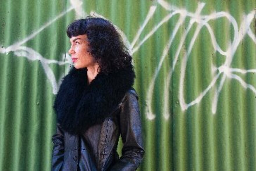 Woman with dark hair standing and wearing a coat with a fur collar, posed against a green corrugated iron fence, looking to the side.